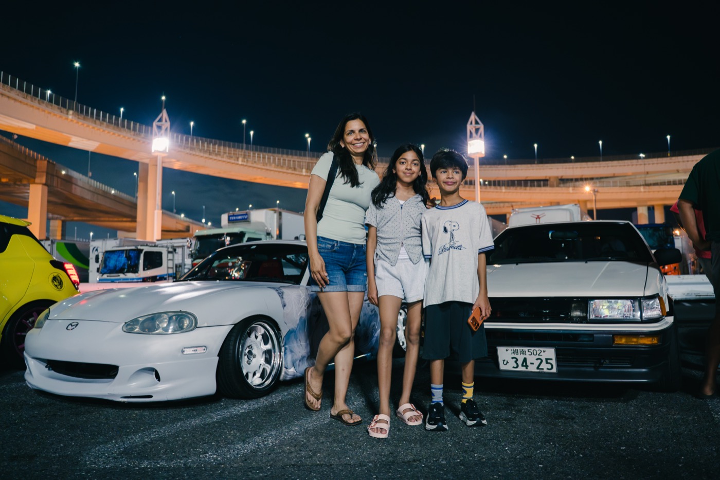 Family on a JDM tour at Daikoku PA with a Toyota AE86 and Mazda Roadster in the evening